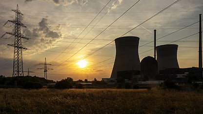 Silueta unei centrale electrice, cu turnuri mari de răcire și linii de transport al energiei care se întind pe cer la apus, văzută de pe un câmp cu iarbă, sub un cer auriu parțial înnorat.